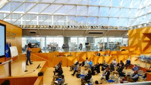 large room with glass ceiling with people sitting in chairs listening to a speaker on stage