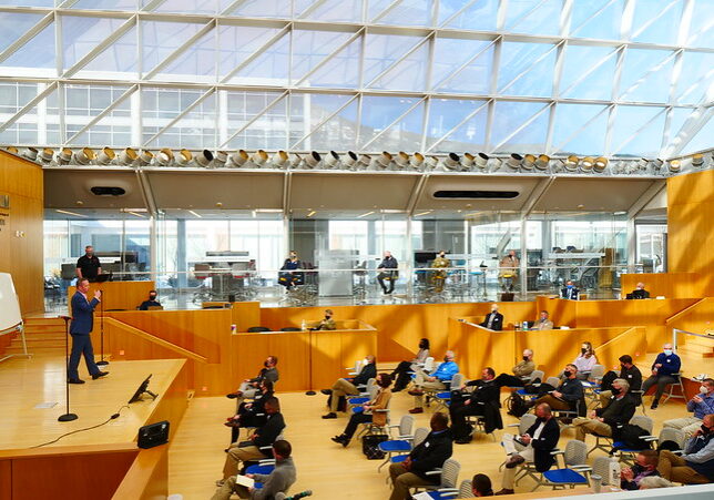 large room with glass ceiling with people sitting in chairs listening to a speaker on stage